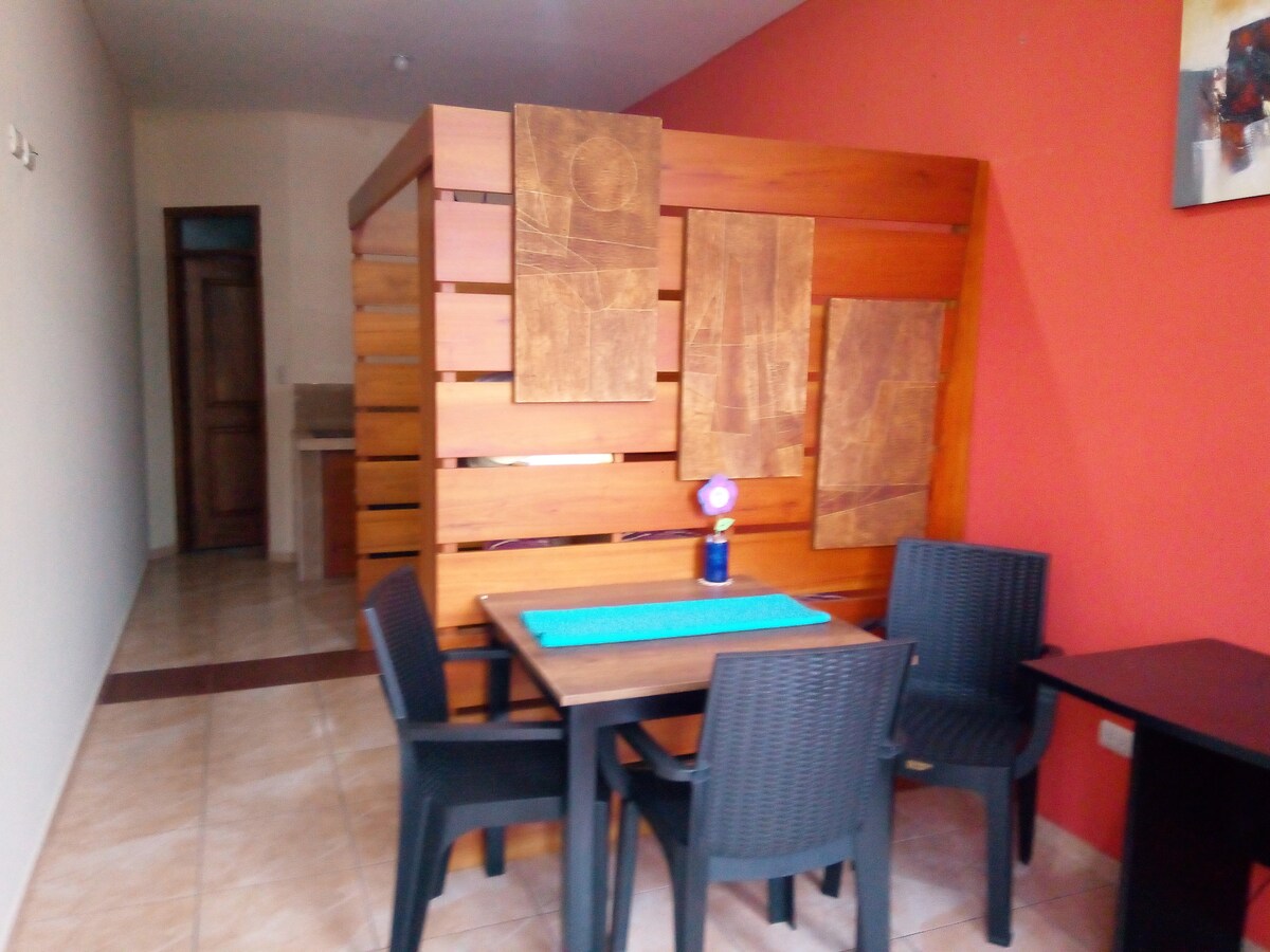 A dining area is presented with a wooden table surrounded by four dark chairs. The backdrop features wooden panels with artistic designs, complemented by a colorful lamp on the table. A muted red wall enhances the space, while a doorway leads to another room.