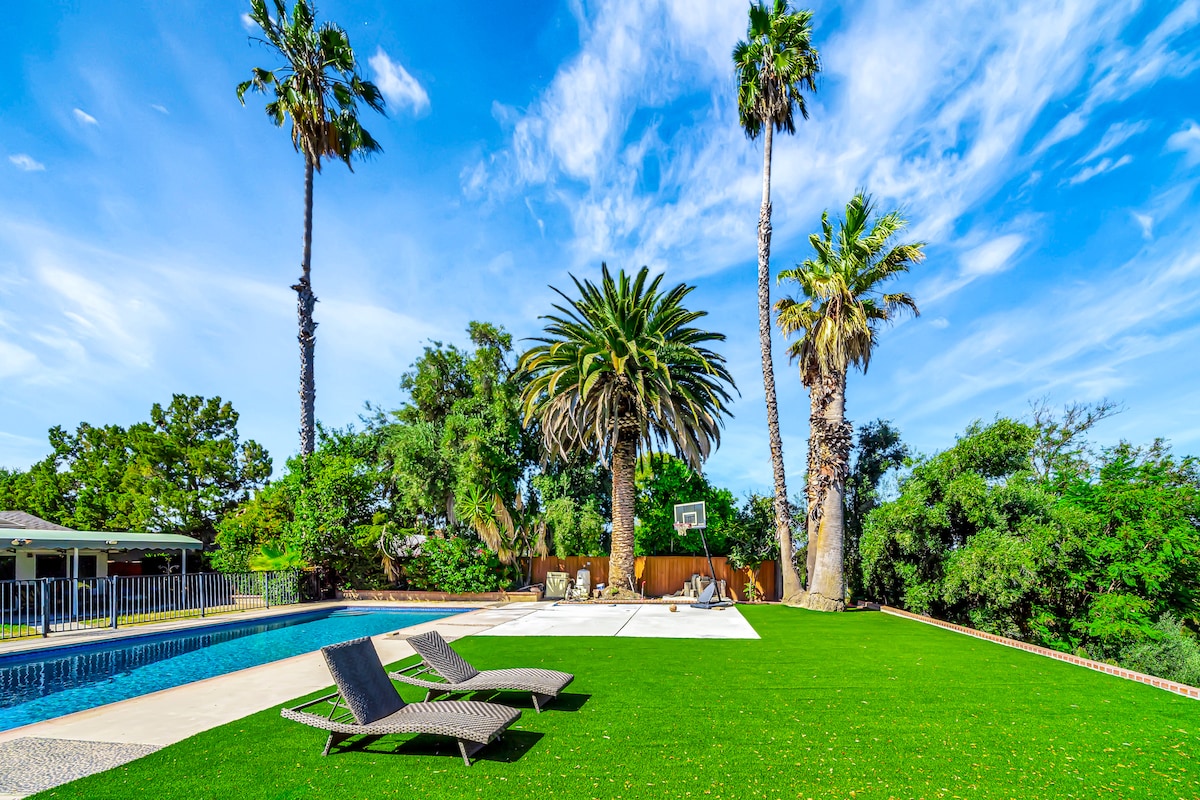 A spacious outdoor area features a large pool surrounded by a lush green lawn. Two lounge chairs are positioned beside the pool, while a basketball hoop stands on a concrete surface. Tall palm trees provide shade and enhance the tropical ambiance.