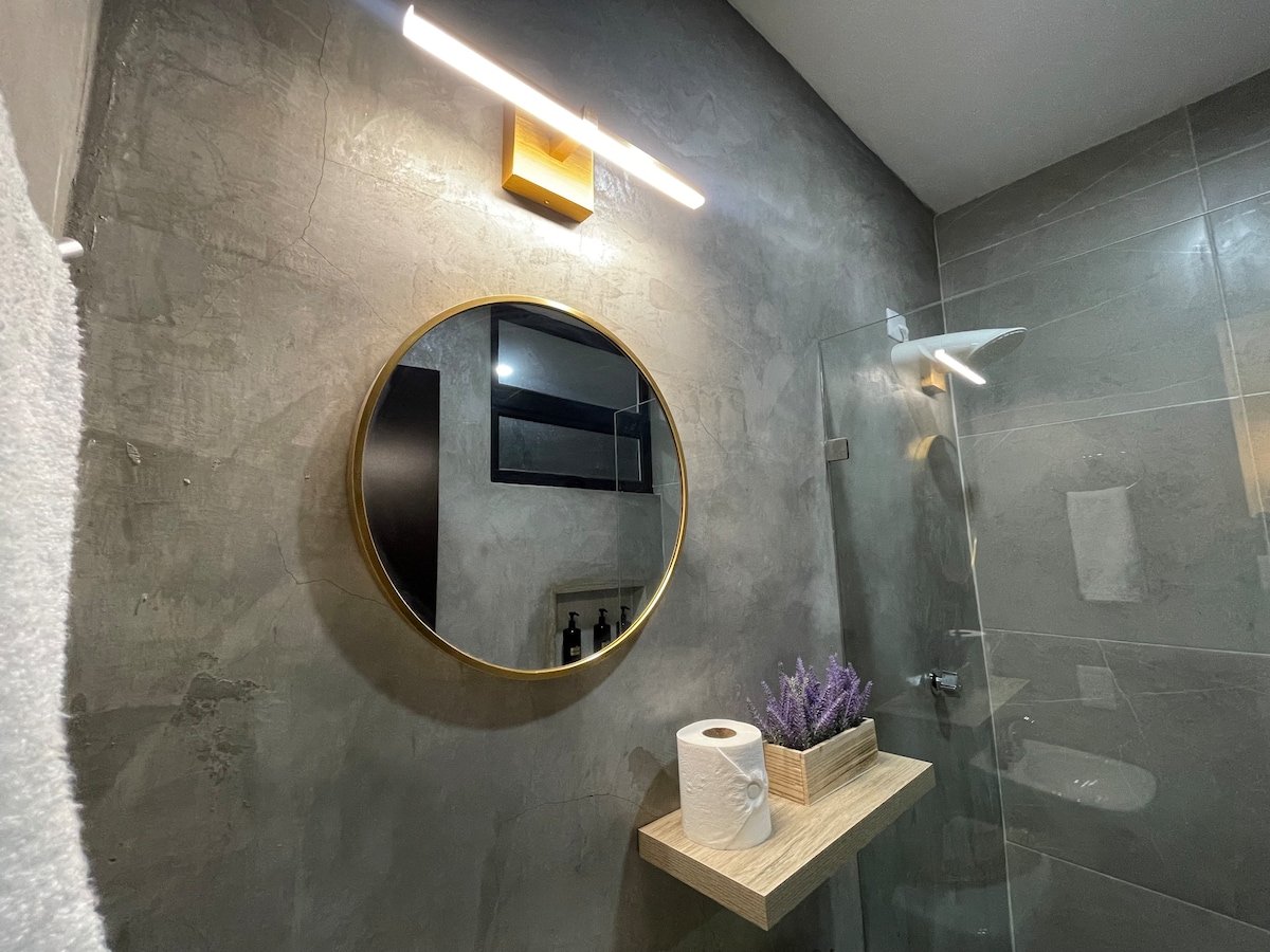 A modern bathroom features a round mirror framed in gold, positioned above a wooden shelf. On the shelf, a roll of toilet paper and a small plant add a touch of decor, while a clear glass shower is visible nearby.