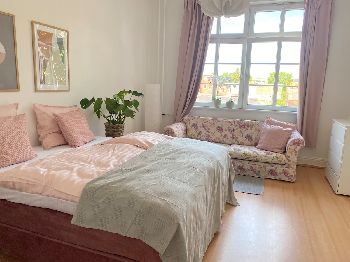 A well-furnished bedroom features a spacious bed with soft linens and decorative pillows. A floral-patterned sofa is positioned near a large window that allows natural light to fill the room. A plant adds a touch of greenery, enhancing the overall ambiance.