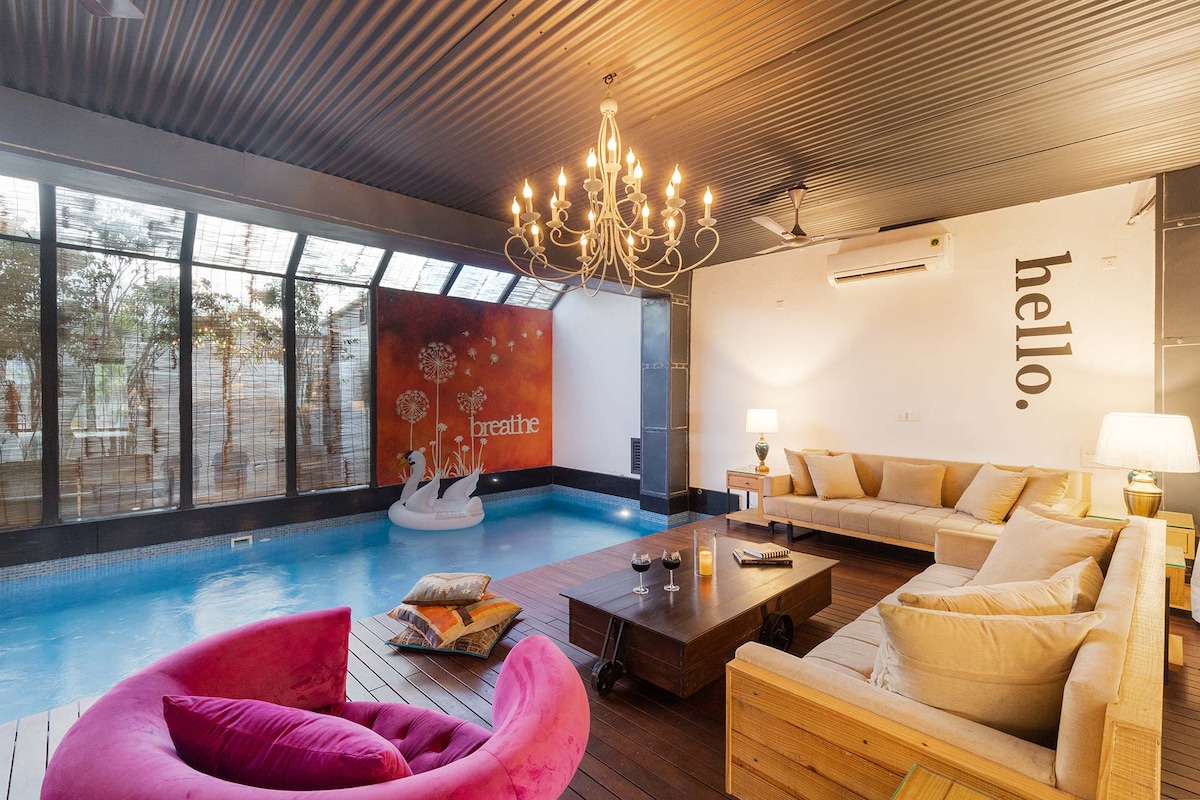 A bright indoor pool area is accentuated by large windows that allow natural light to fill the space. A comfortable beige sofa and vibrant pink seating create an inviting atmosphere. A decorative chandelier hangs above, complementing the modern decor, while artwork adorns the walls.