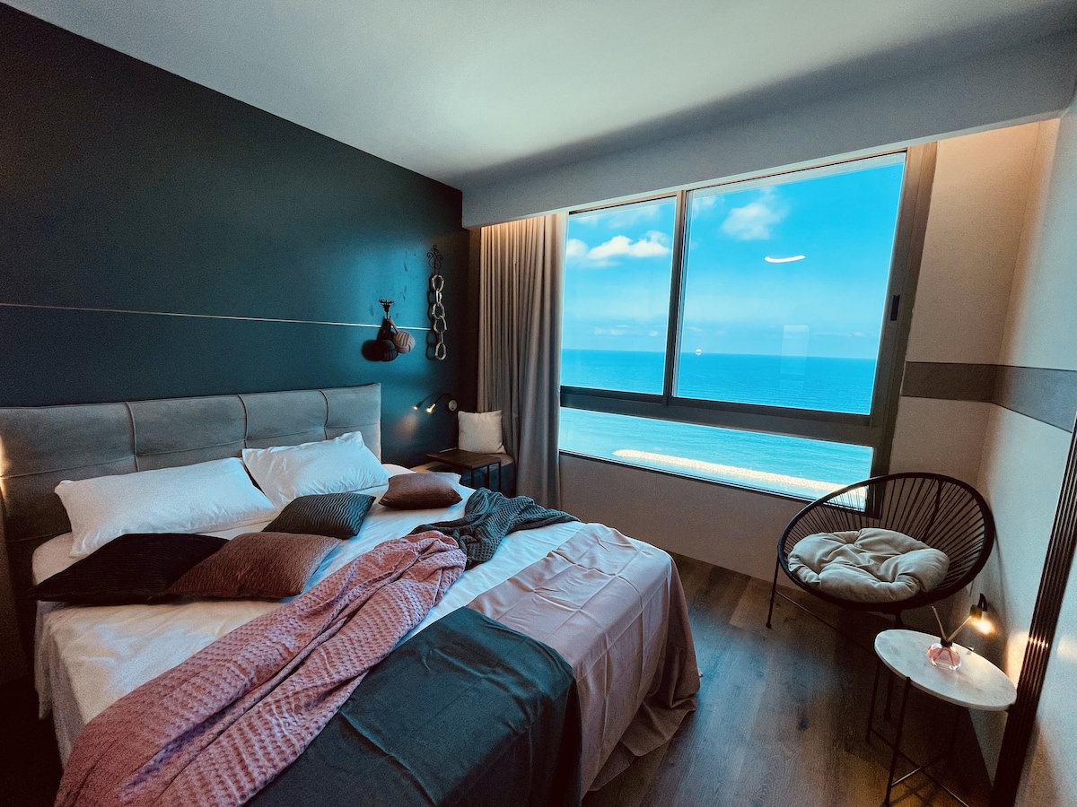 A spacious sleeping area features a well-made bed with subtle bedding tones. A large window offers a view of the ocean, with light filtering through sheer curtains. A small side table displays a cozy blanket, enhancing the tranquil setting.