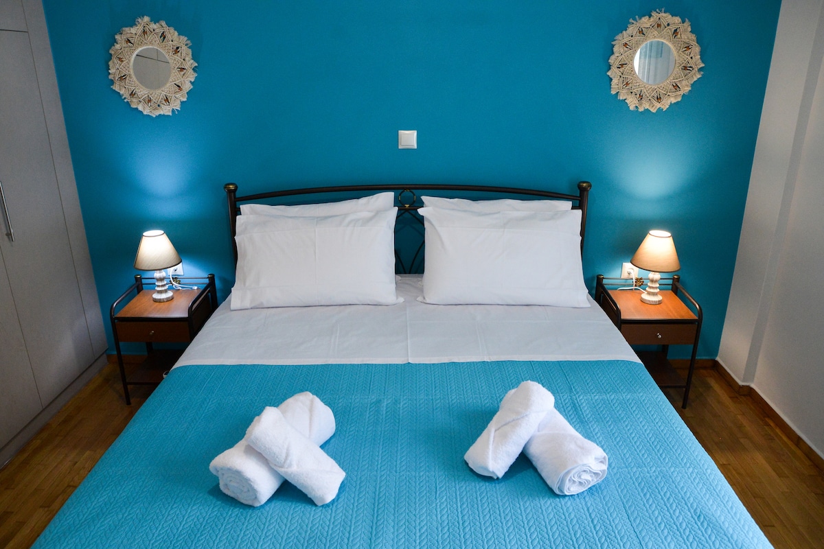 A bright sleeping area features a spacious bed with white linens and a light blue blanket. Two decorative lamps with warm light are placed on nightstands beside the bed. The walls are painted a vibrant blue, complemented by two round mirrors.