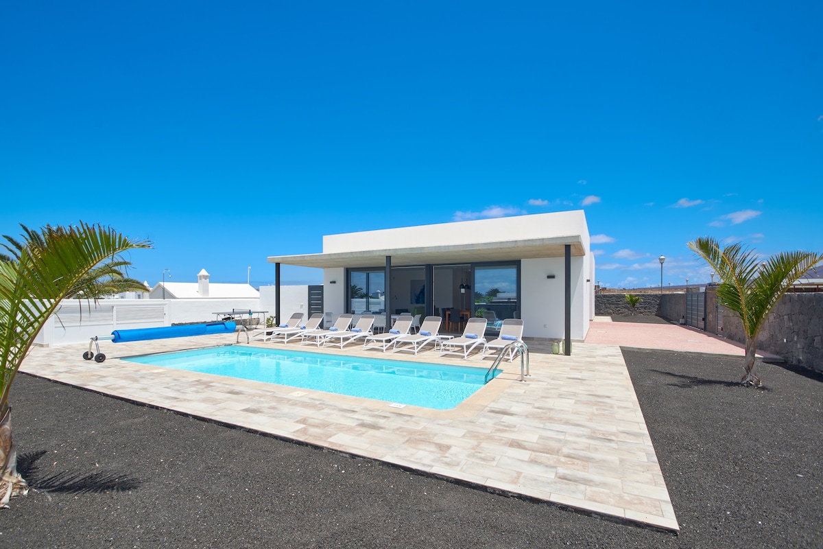 A modern villa is shown beside a private pool, surrounded by a spacious patio. Several lounge chairs are positioned along the poolside. A clear blue sky complements the clean lines and contemporary design of the villa, which features large windows for ample light.