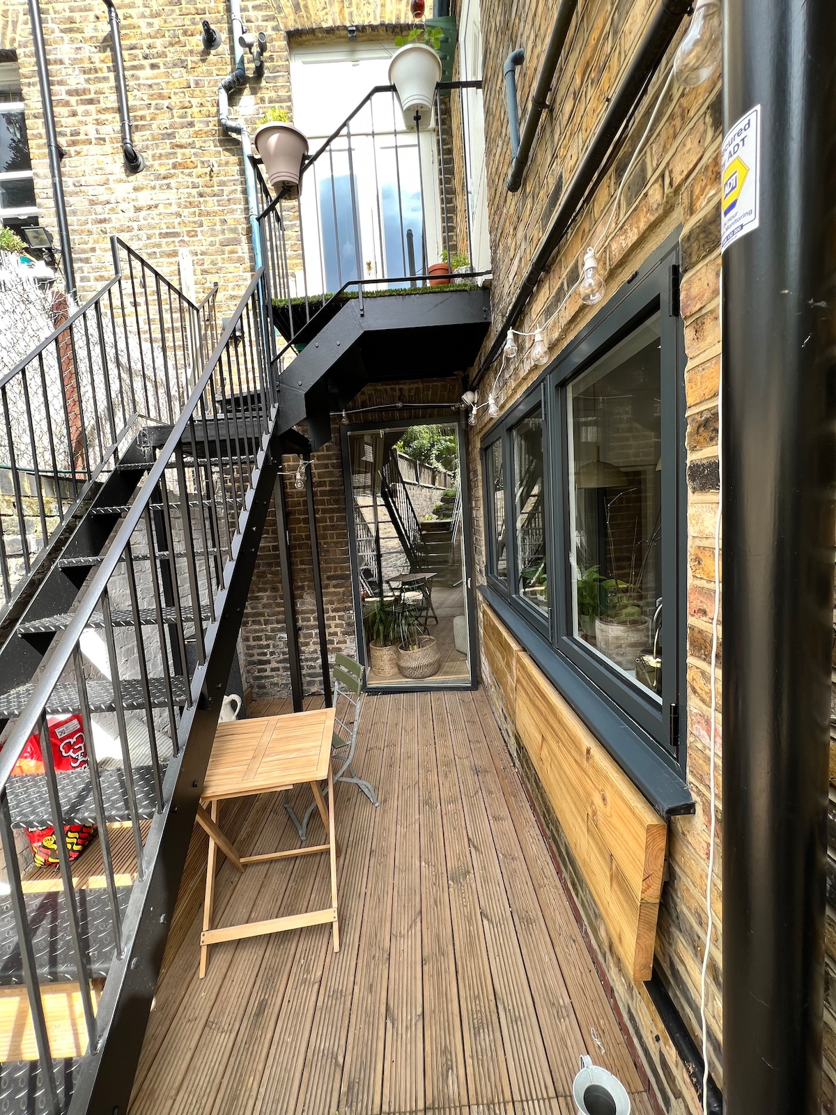 A wooden deck extends along the exterior of the flat, bordered by a rustic stone wall. A small table with a chair is positioned against the wall, providing a cozy outdoor spot. Stairs lead up to a higher level, while a glass door opens into the indoor space.