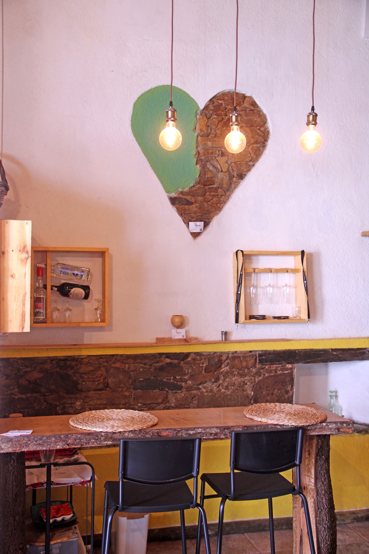 A cozy bar area is showcased with a natural wood countertop and two black chairs. Above, three hanging light bulbs illuminate a heart-shaped stone feature on the white wall, complemented by a wooden shelf displaying glassware and bottles.