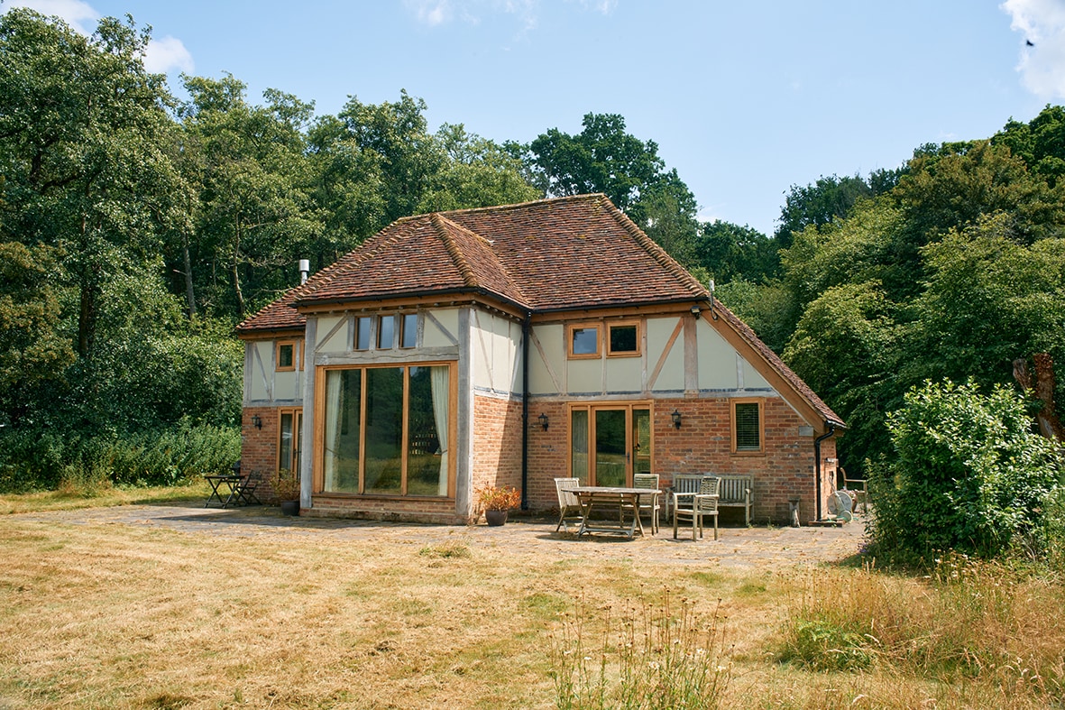 A charming exterior of the property is highlighted, featuring a combination of brick and timber framing. Large windows allow natural light to fill the indoor spaces, while outdoor seating is arranged on a grassy area, surrounded by lush greenery.