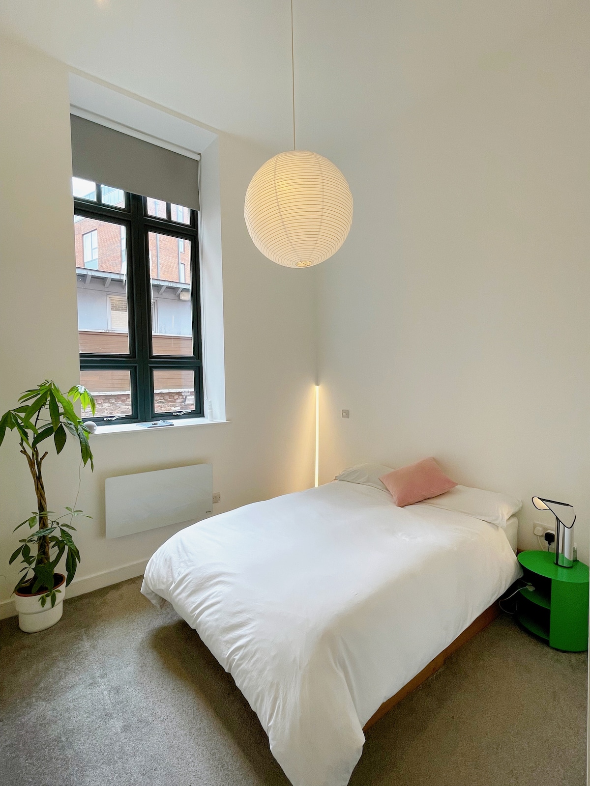 A bright bedroom features a spacious bed with crisp white bedding and a pink accent pillow. A modern pendant light provides soft illumination. A green nightstand is positioned beside the bed, along with a small bedside lamp. Large windows allow natural light to fill the room.