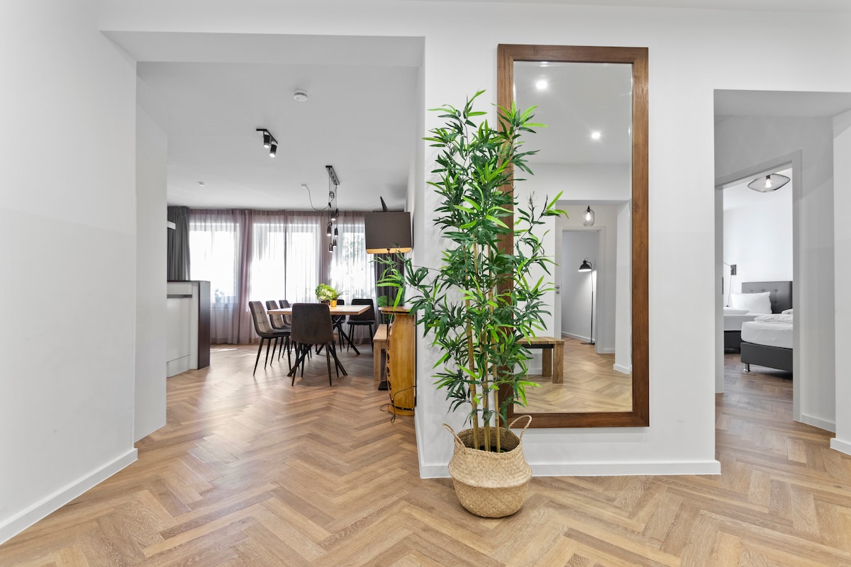 A bright entryway features stylish herringbone flooring and a decorative plant in a woven basket. A large mirror reflects the space and leads to an inviting dining area with a wooden table. Soft natural light filters through sheer curtains, enhancing the welcoming ambiance.