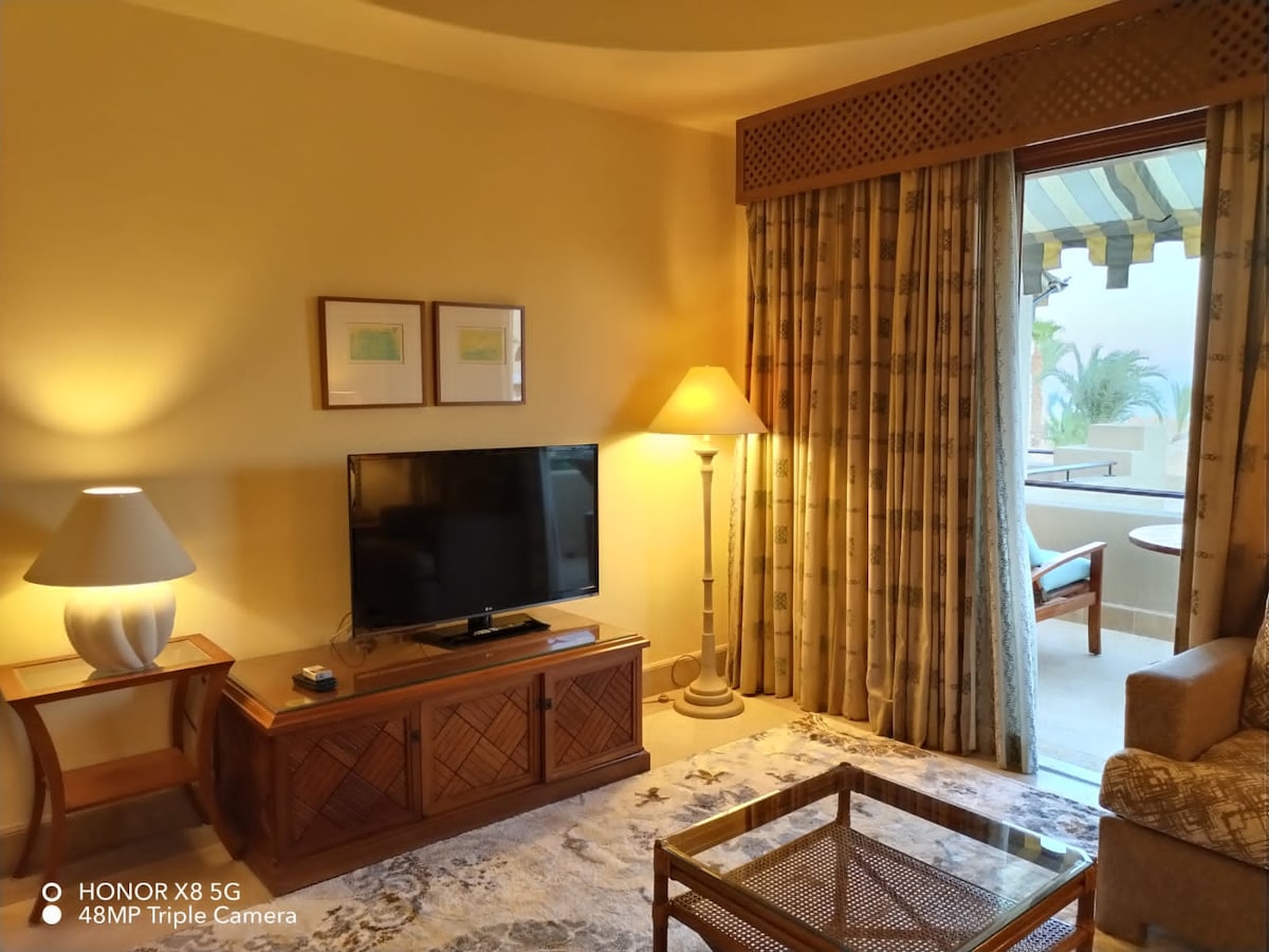 A comfortable living area includes a flat-screen television mounted on a wooden cabinet. A glass coffee table sits on a patterned rug, and large curtains frame a window that allows natural light to fill the space. A soft lamp provides additional illumination.