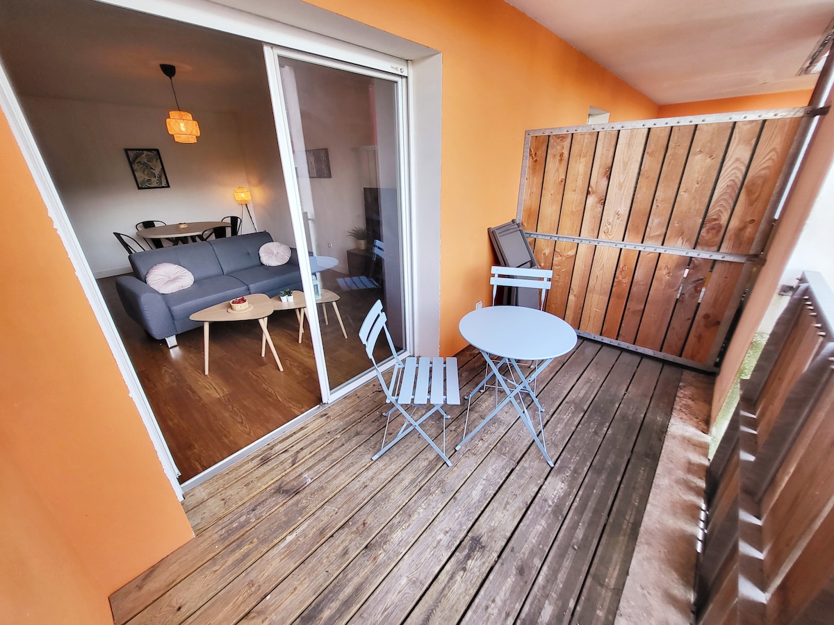 A small balcony is shown, featuring a wooden deck and a light blue table with matching chairs. The walls are painted in a warm orange hue, contrasting with the natural wood. Beyond the glass doors, a living space is visible, creating a seamless indoor-outdoor connection.