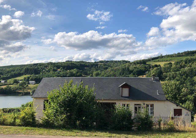 A serene countryside house is nestled along the edge of a lake, surrounded by lush greenery and rolling hills. A sloped roof and multiple windows provide a sense of openness, while a tranquil sky with scattered clouds enhances the peaceful setting.