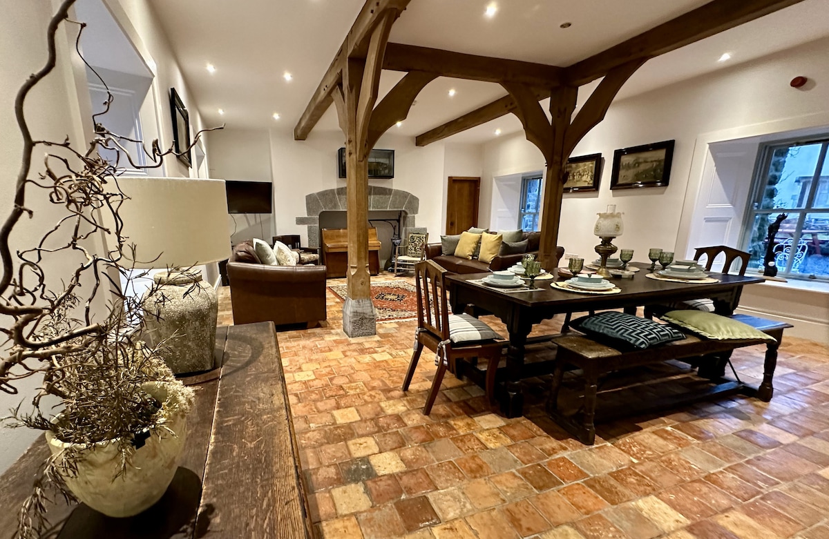 An inviting sitting area features exposed wooden beams and a large dining table, surrounded by comfortable seating. A stone fireplace serves as a focal point, with soft lighting illuminating the warm terracotta floor. Decorative items add subtle texture and interest to the space.