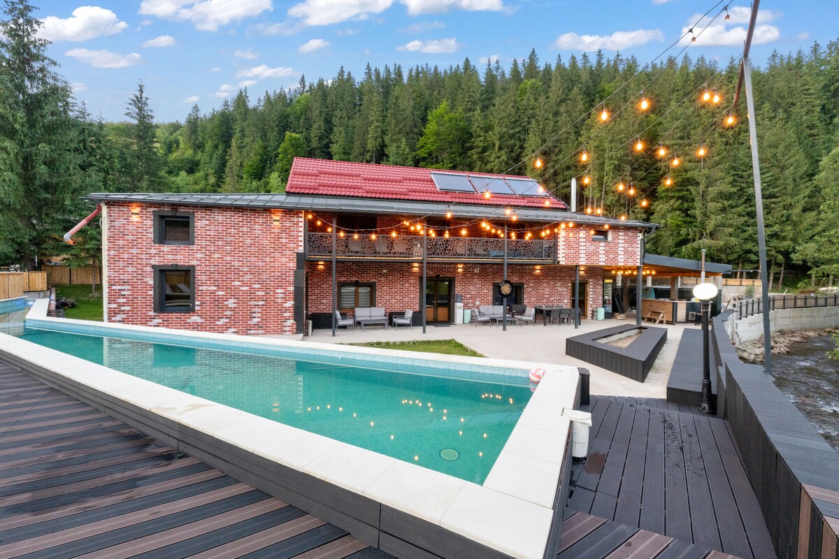 A charming brick building is set against a backdrop of lush greenery. A sparkling swimming pool is bordered by a wooden deck, with lounge seating positioned nearby. Soft outdoor lighting enhances the inviting ambiance in the evening, complementing the scenic landscape.
