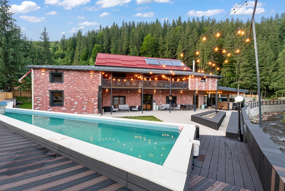 A contemporary brick house is nestled among tall trees, featuring a spacious outdoor area with a swimming pool. String lights illuminate the patio, enhancing the inviting atmosphere. Large windows reflect the surrounding greenery, complementing the natural setting.