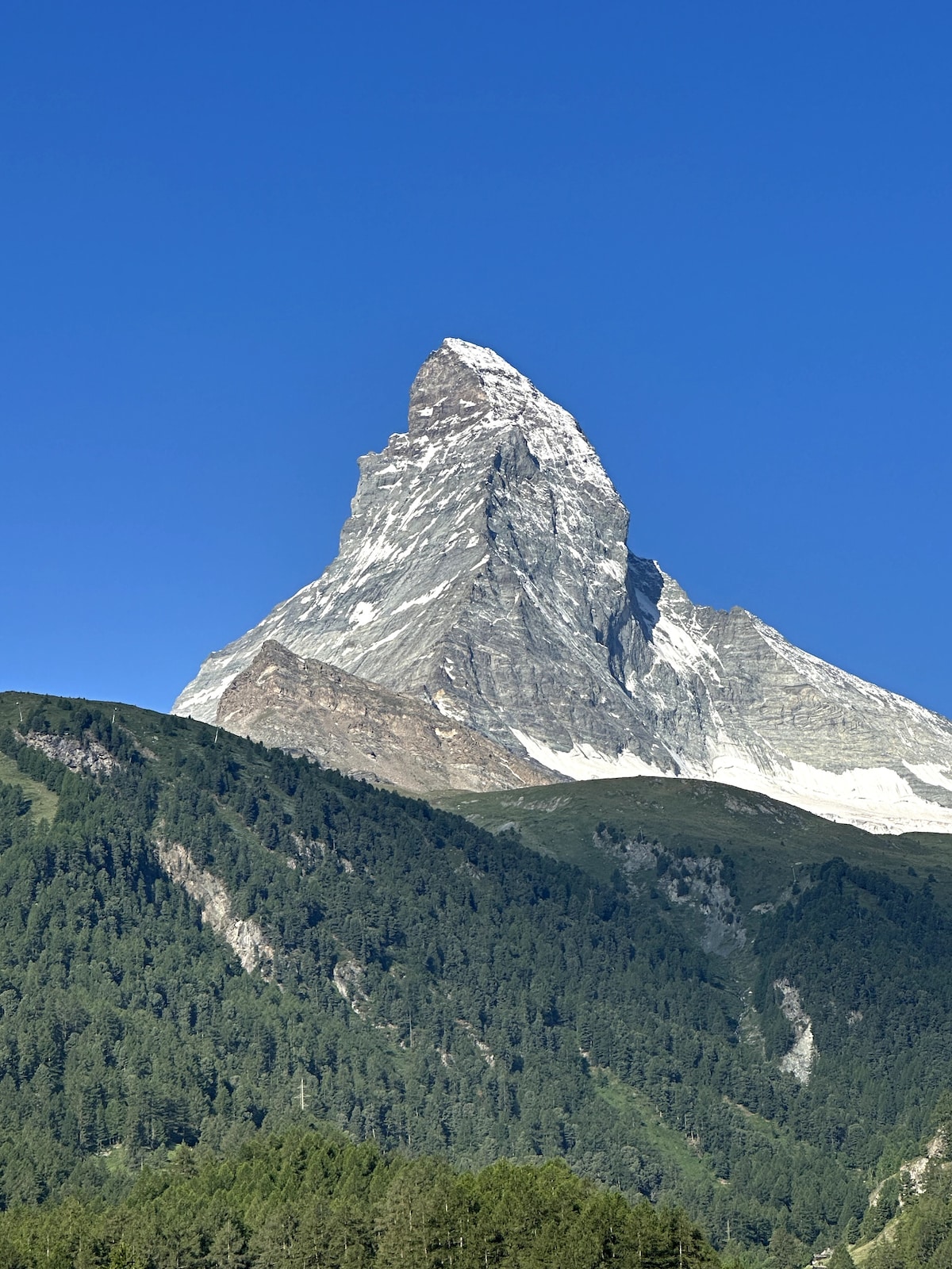 A prominent mountain peak is captured against a clear blue sky. The mountain, displaying a distinctive pyramid shape, is partially covered with snow at the summit, while lush green forests are visible at its base. This serene landscape embodies natural beauty.