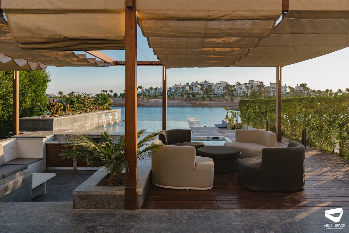 An outdoor lounge area features comfortable seating arranged around a central table. Natural greenery surrounds the space, with a view of the lagoon in the background. The setting is designed to blend with the tranquil landscape, providing an inviting space for relaxation.