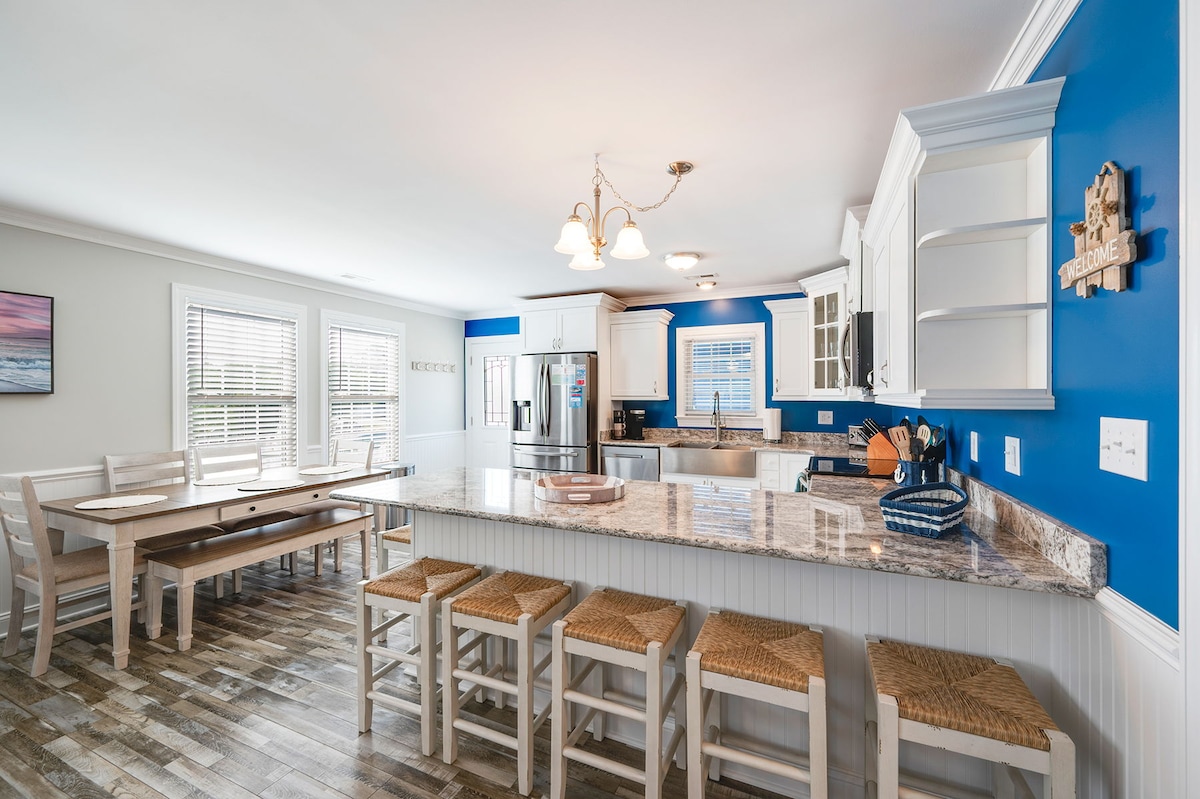 A bright and airy kitchen and dining area features a large granite island with seating for several guests. Light wood stools surround the island, and a spacious farmhouse table is positioned nearby. The room is accented with blue walls and natural light streaming through windows.