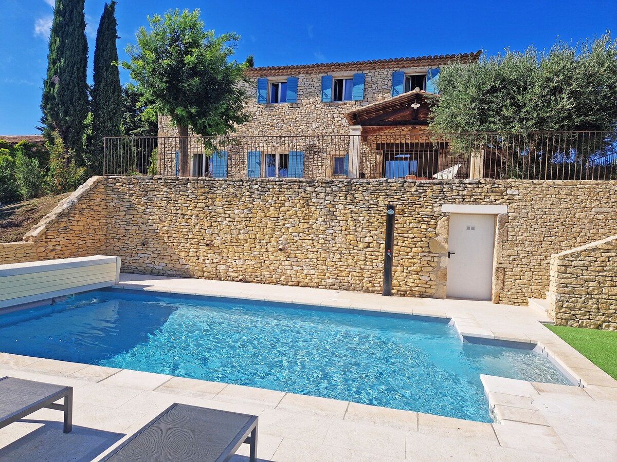A clear blue swimming pool reflects sunlight, surrounded by a stone wall and lush greenery. The property features a two-story structure with large windows and a wooden entrance visible in the background, complemented by a sunbathing area with minimalist lounge chairs.