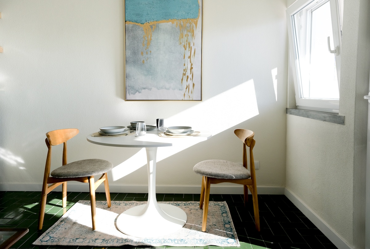 A round dining table with a white surface is positioned under a painting featuring shades of blue and gold. Two wooden chairs with cushioned seats are arranged around the table. Bright light streams through a nearby window, creating a warm atmosphere in the space.