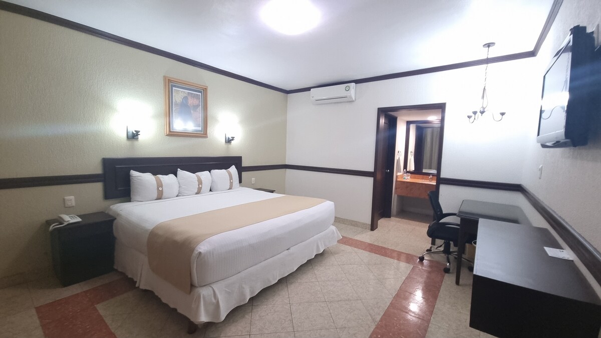 A spacious guest room features a large bed with crisp white linens and neutral accents. Soft lighting from wall sconces provides illumination. A desk and chair are positioned near a flat-screen TV. The adjoining bathroom can be seen through an open doorway.