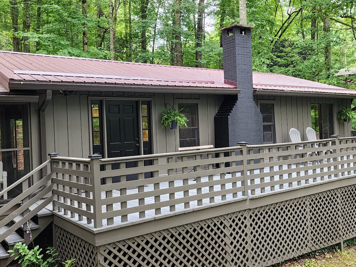 The cabin features a large front deck surrounded by lush greenery. A black chimney stands prominently at one end, with two chairs placed for seating. The entrance door is framed by potted plants, adding a touch of nature to the welcoming facade.