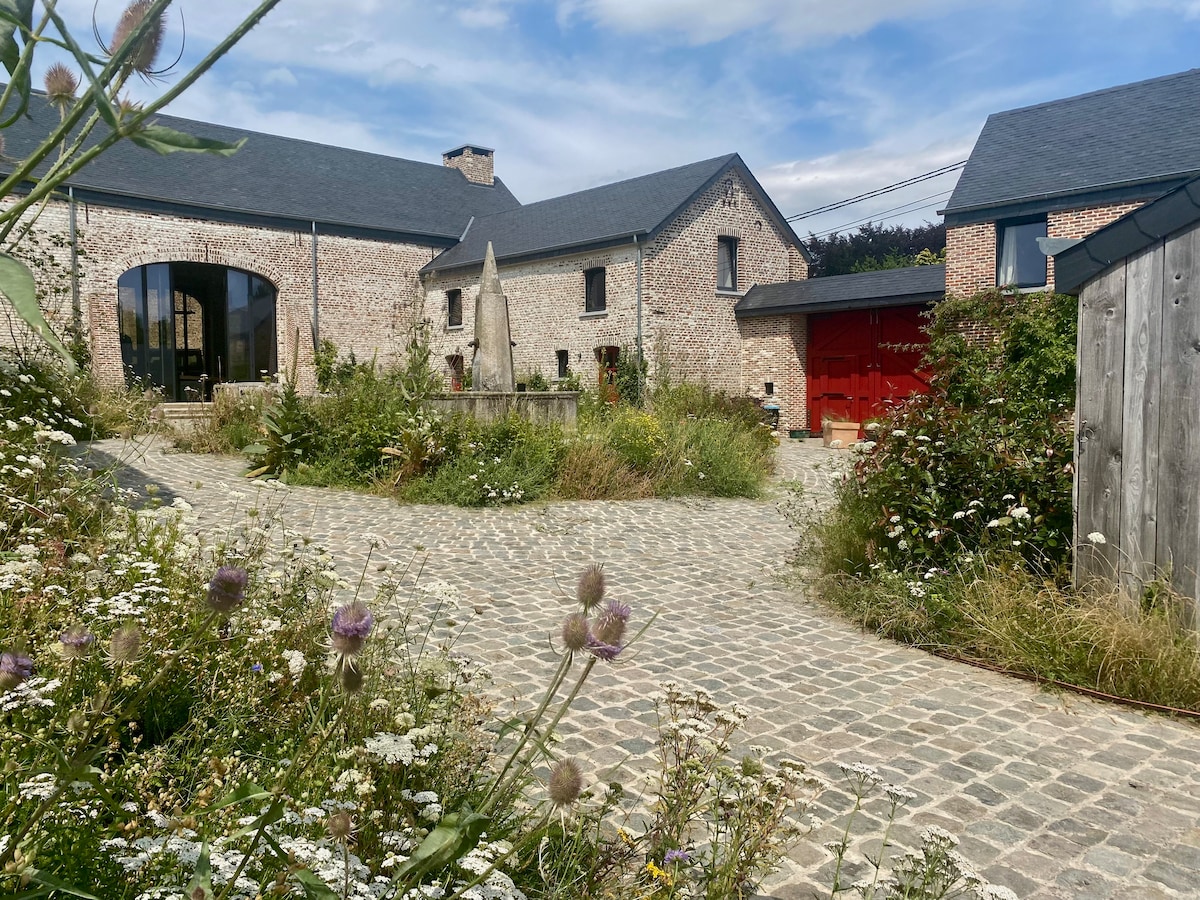 A cobblestone path meanders through a natural garden filled with wildflowers, leading towards charming brick buildings. A central fountain is visible, surrounded by greenery, with a backdrop of rustic structures showcasing red accents. The scene reflects a serene and countryside atmosphere.