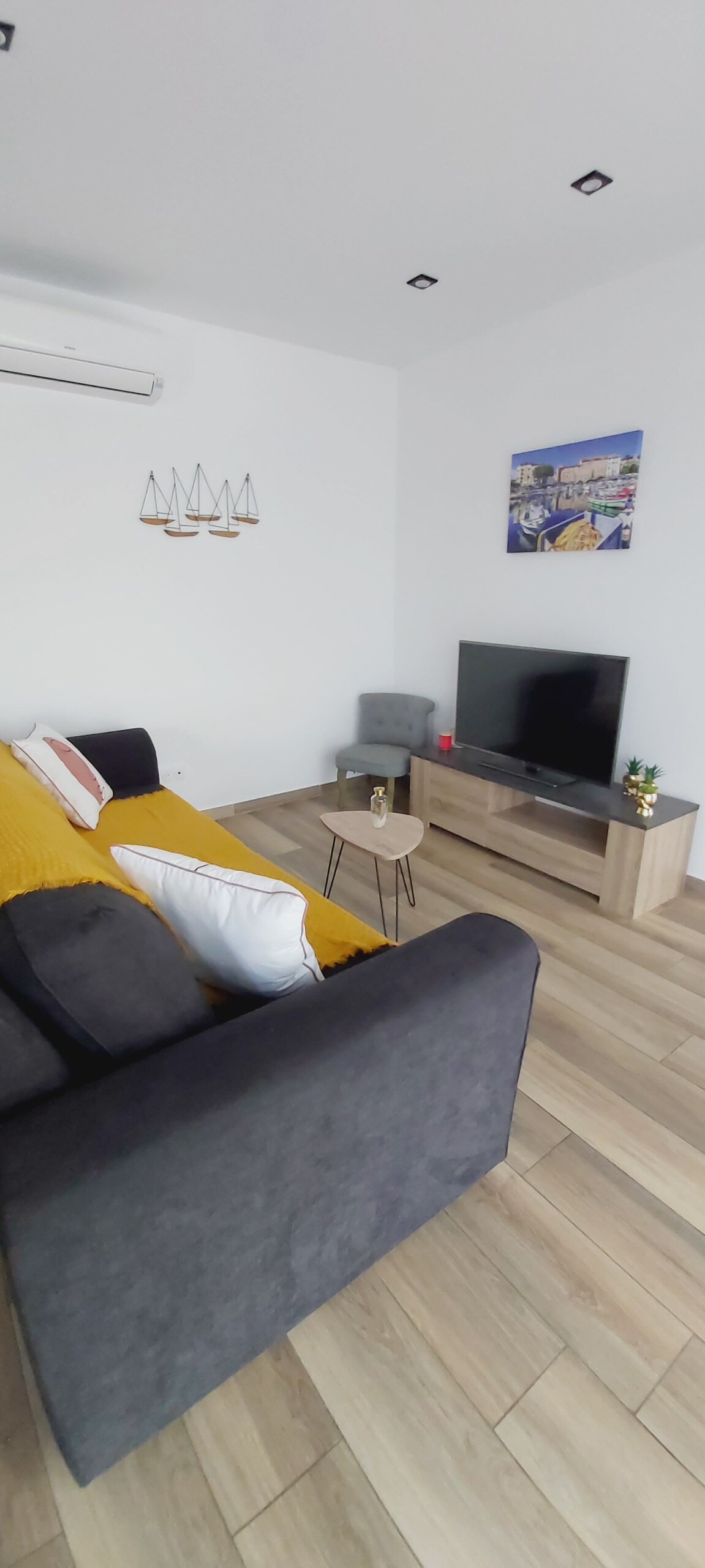 A welcoming living area is featured with a dark-colored sofa adorned with cushions and a bright yellow throw. A small side table sits next to the sofa, while a television is placed on a light wood media console. Light wood flooring enhances the space.