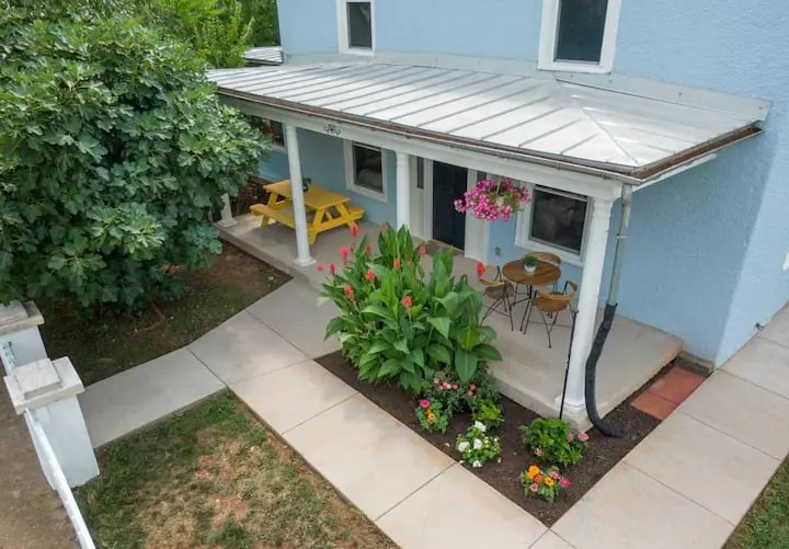 A welcoming front porch is showcased, featuring a yellow picnic table and two small round chairs. Colorful potted flowers and lush greenery frame the entrance, while a neatly paved pathway leads to the house, enhancing the inviting atmosphere of the exterior.