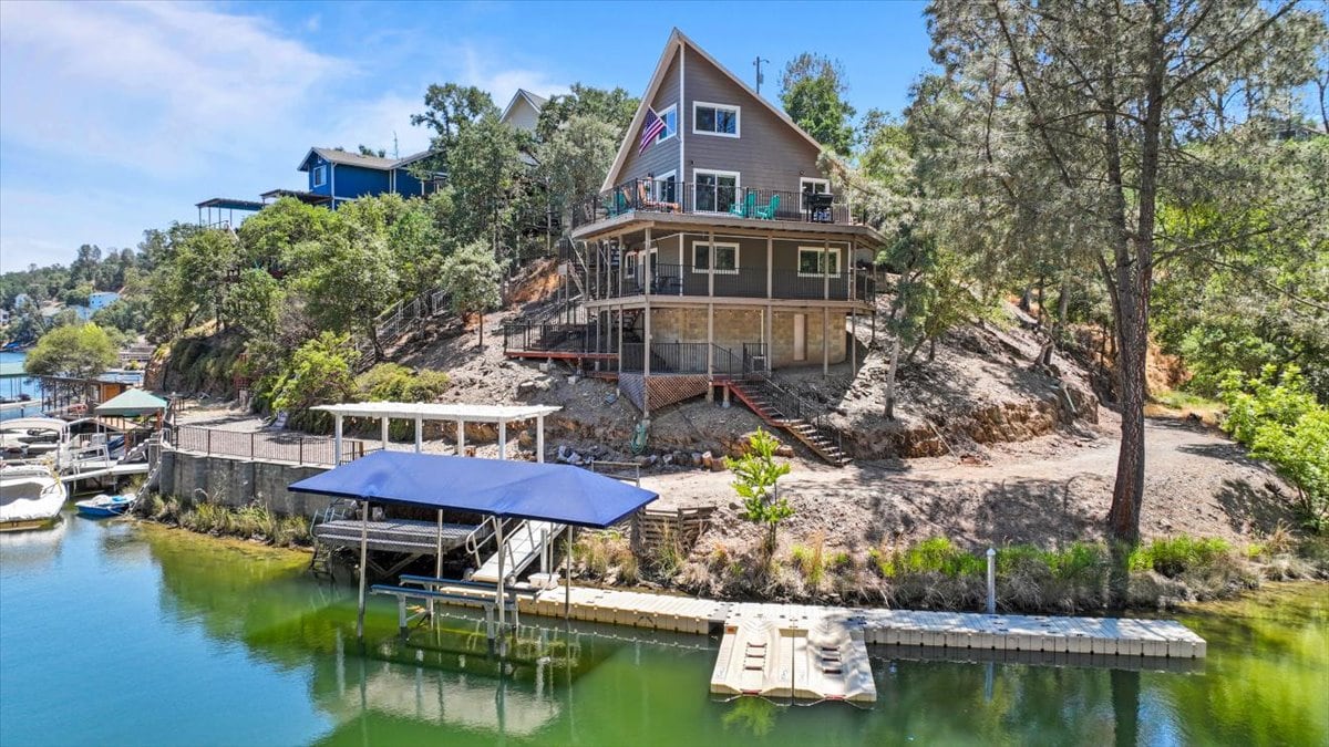A waterfront home is positioned on a hillside, featuring multiple levels with large windows. A private dock is visible along the water, with a shaded area beneath a canvas canopy. Surrounding foliage adds greenery, emphasizing the serene lakeside setting.
