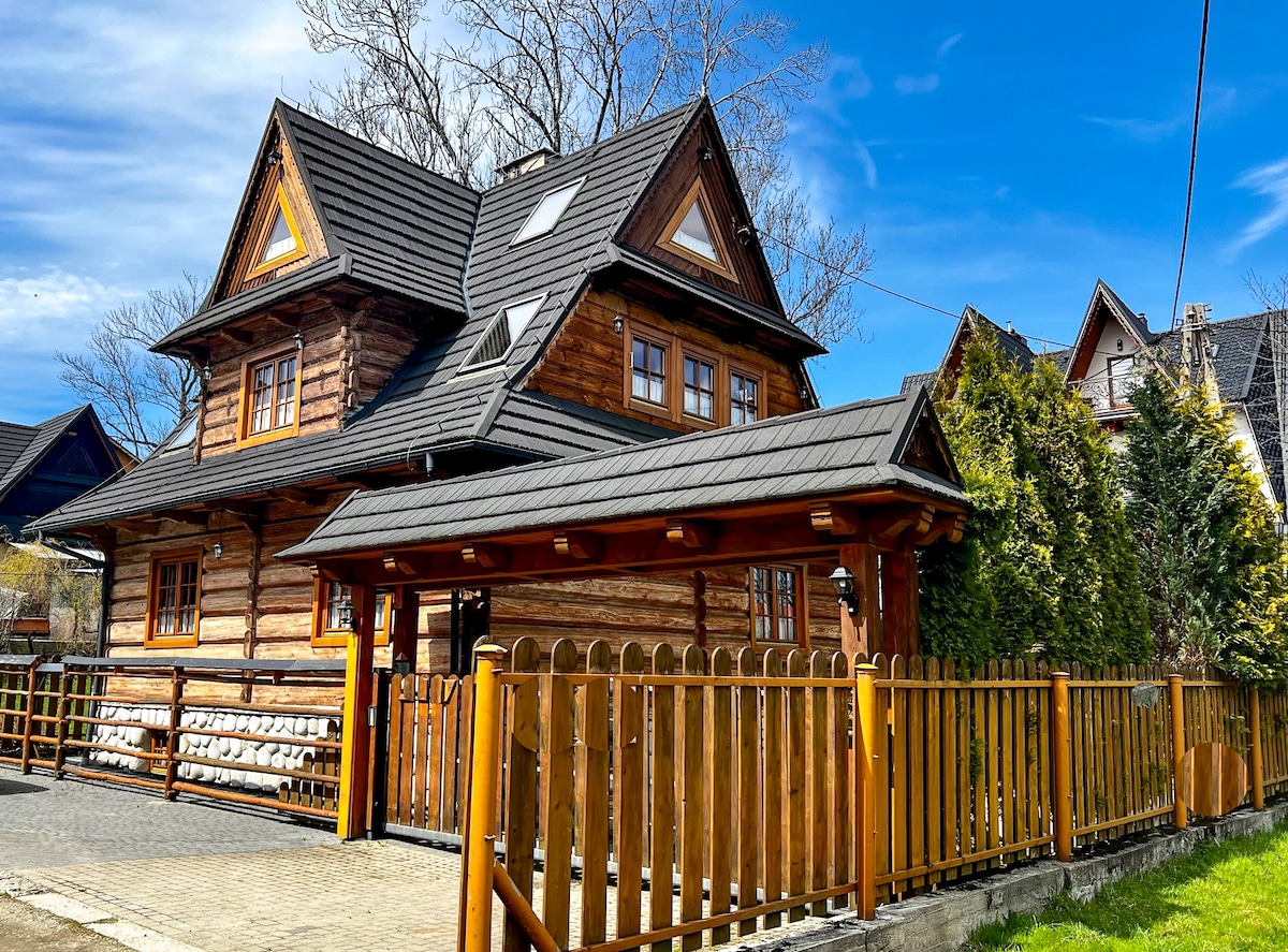 The wooden house is characterized by a traditional alpine design, featuring a steeply pitched roof and decorative gables. A charming wooden fence encircles the property, and tall trees provide privacy. The exterior showcases natural wood tones against a clear blue sky.