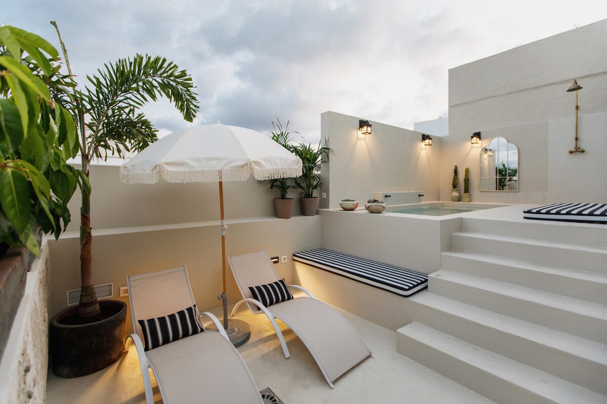 A spacious rooftop terrace features lounge chairs with striped cushions and a shaded umbrella. Plants are arranged in decorative pots, and a small private pool is visible on an elevated surface, surrounded by elegant decor and soft lighting for a relaxing ambiance.