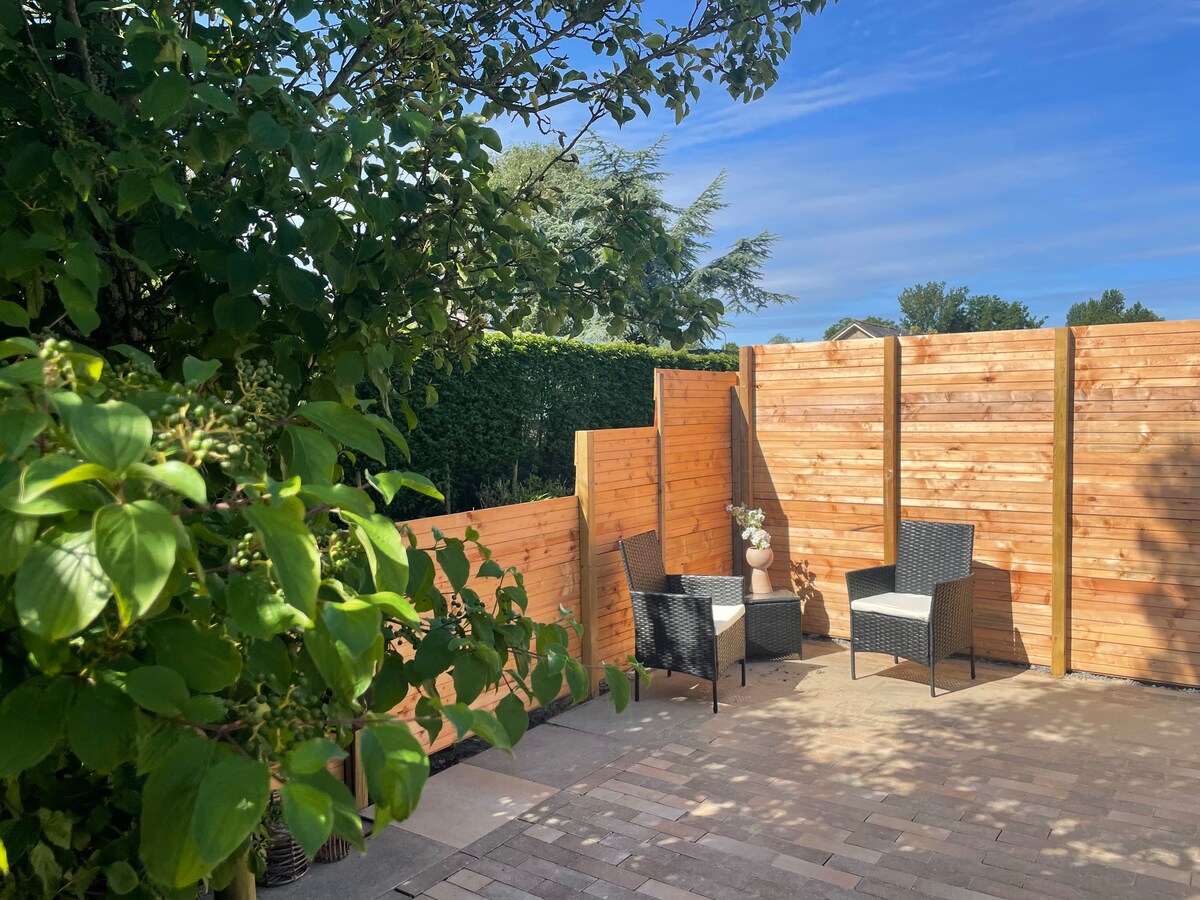 A private garden area is enclosed by wooden fencing, featuring two woven chairs positioned on a stone-paved surface. Lush greenery is visible in the foreground, with clear blue skies above, creating a serene outdoor retreat.