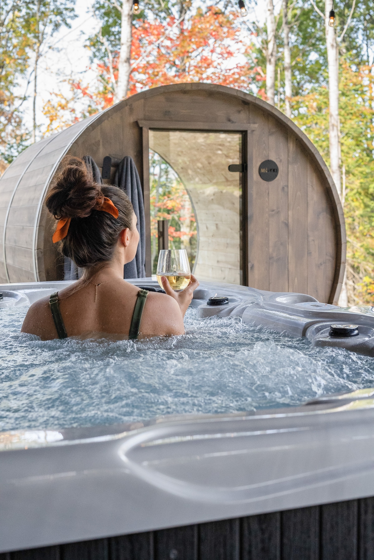 A person is seen relaxing in a bubbling spa, holding a glass of wine. The background features a wooden sauna structure with a rounded design, framed by colorful autumn foliage. Natural light creates a warm ambiance through the windows.
