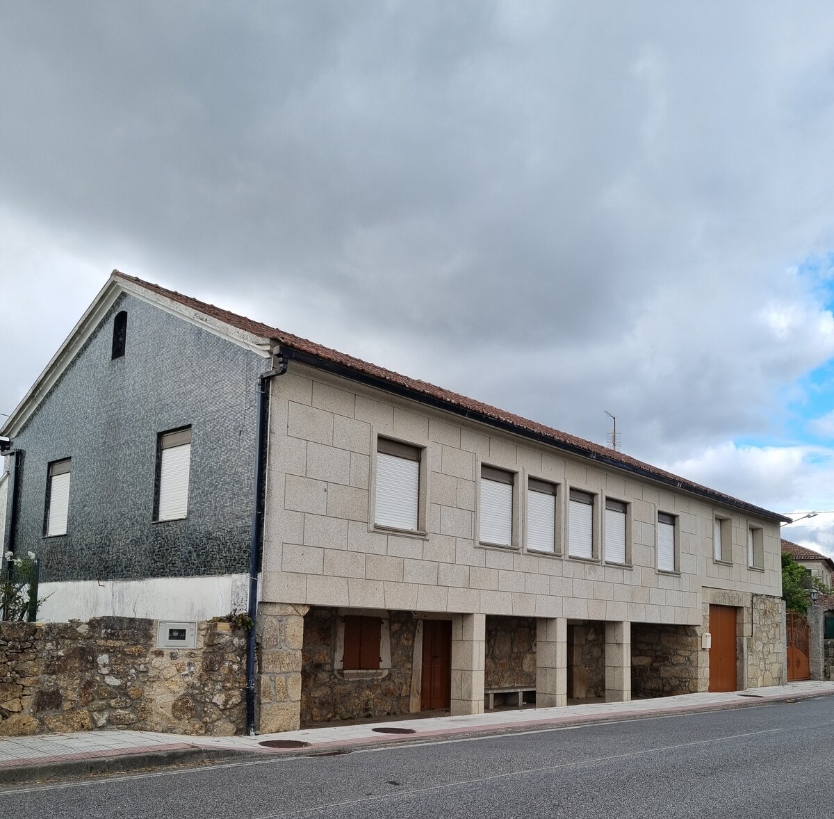 The exterior of the property features a two-story building with a combination of stone and modern siding. Windows are uniformly arranged, providing a balanced look. A smooth road runs alongside the building, with a cloudy sky overhead, creating a calm rural setting.