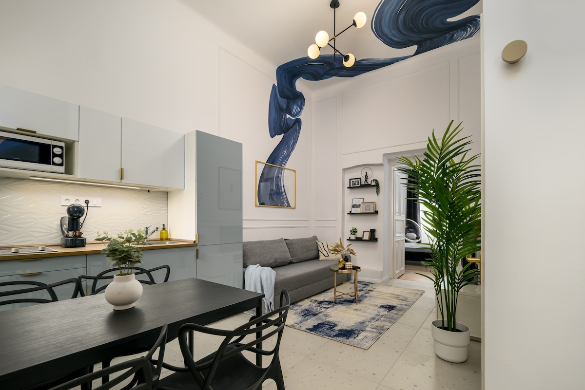 A bright and modern living space features a sleek kitchen area complemented by a wooden dining table and black chairs. A cozy seating area is highlighted by a gray sofa, with a colorful wall mural adding an artistic touch. Indoor plants enhance the lively atmosphere.