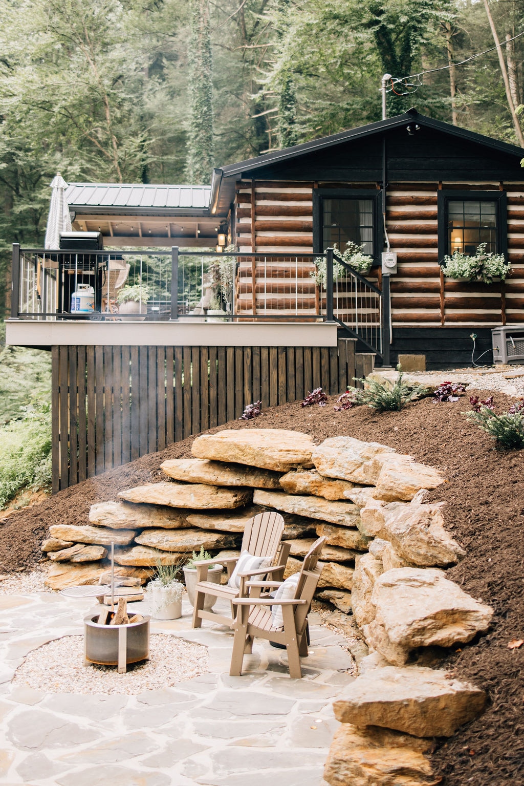 A cozy seating area is situated on a flagstone patio, featuring two adirondack chairs positioned around a fire ring. The cabin is visible above, surrounded by lush greenery, while flower beds add color to the natural landscape.