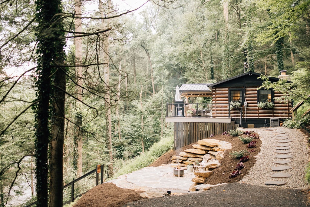 The exterior view of the log cabin is set amidst a lush, wooded landscape. A stone path leads to the cabin, which features a spacious outdoor deck adorned with plants and seating. The serene ambiance of the surrounding nature invites relaxation.