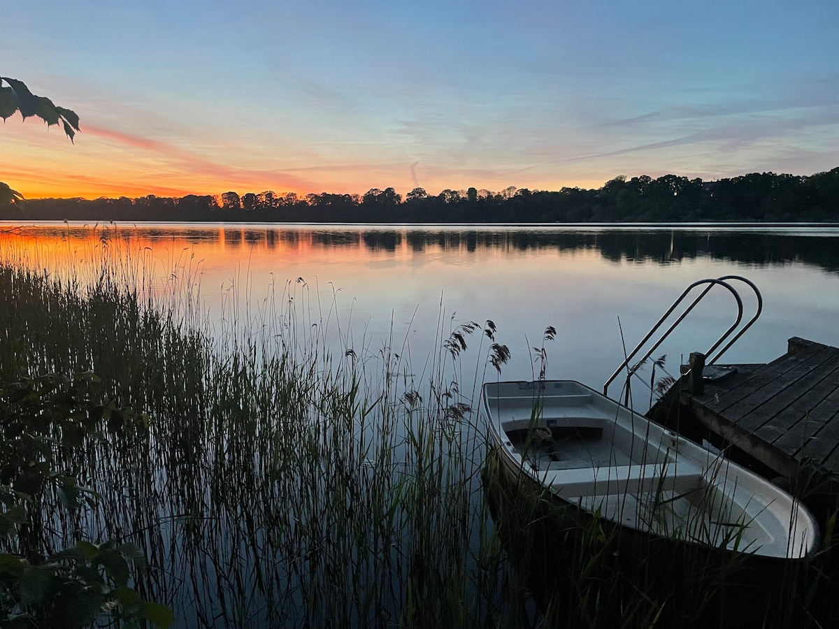 A serene lakeside scene is captured at sunset, showcasing calm waters reflecting vibrant hues of orange and purple. A small rowboat is tied to a wooden dock, surrounded by tall grasses and framed by silhouettes of trees lining the shore.