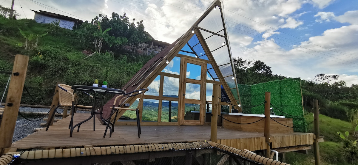A contemporary A-frame structure is situated on a wooden deck, showcasing large glass doors that provide a view of the surrounding landscape. A comfortable seating area is present, with a table and chairs, and a private jacuzzi can be seen on the deck.