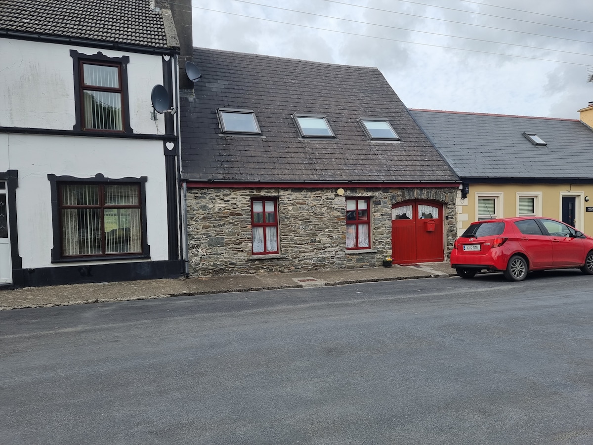 The exterior of the coach house features a traditional stone façade with a distinctive red door. Windows adorned with white curtains allow for natural light. A red car is parked in front, and neighboring structures showcase a blend of textures and colors, enhancing the neighborhood's character.