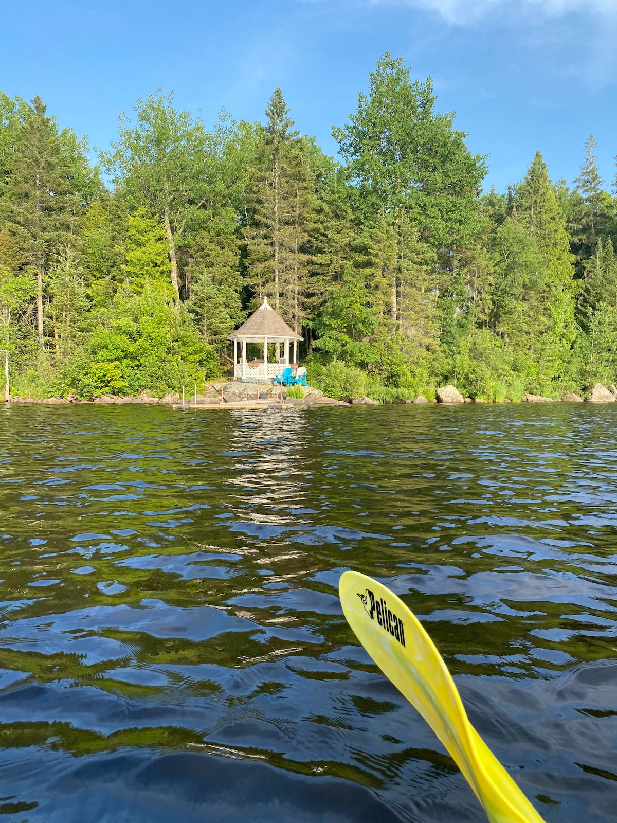 A serene lakeside view is captured from the water, featuring a gazebo nestled among lush trees. The calm water reflects the vibrant greenery, and a glimpse of seating is visible on the gazebo's deck, inviting relaxing moments by the lake.