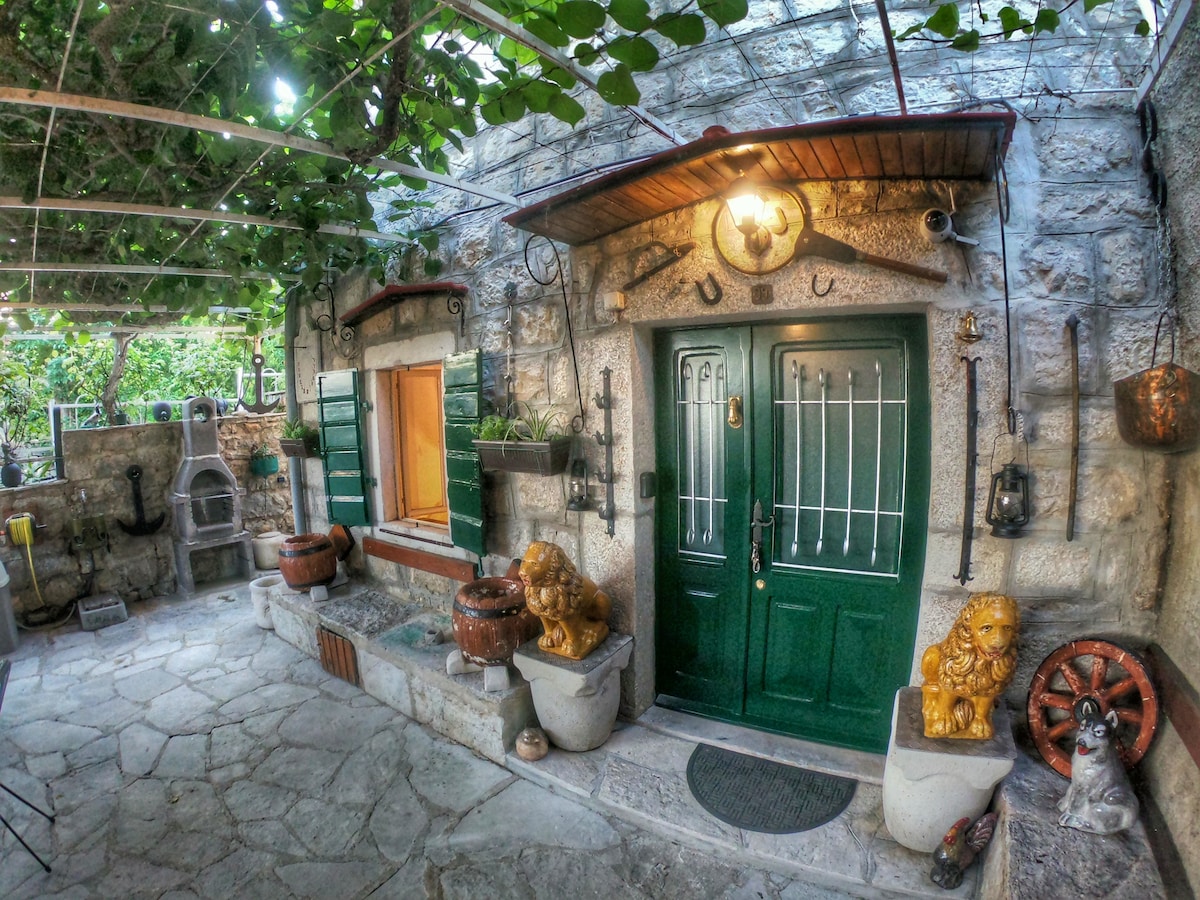 The entrance features a pair of green double doors framed by stone walls, reflecting traditional architecture. An array of decorative pots and rustic artifacts is displayed around the doorway, while a canopy of grapevines provides gentle shade over the outdoor area.