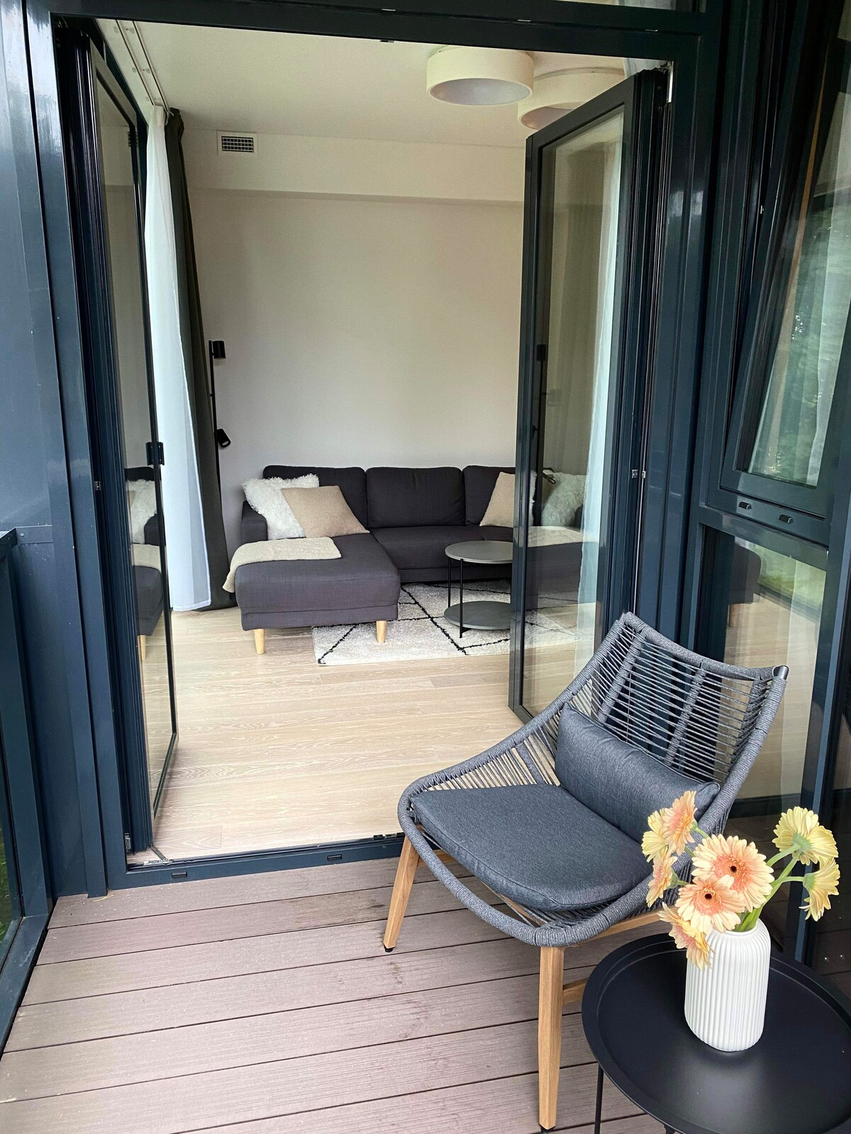 A private balcony is depicted, featuring a comfortable chair and a small side table with a vase of vibrant flowers. Large glass doors lead to a living area with a cozy sofa and a coffee table, merging outdoor and indoor spaces.