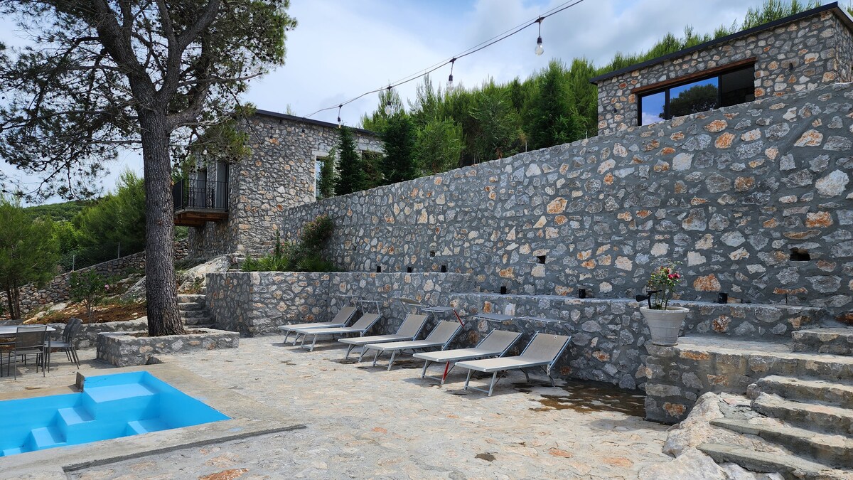 A sun-drenched outdoor area features a modern pool surrounded by stone paths. Several lounge chairs are arranged for relaxation, and a stone wall adds character to the space. Lush greenery can be seen in the background, enhancing the natural setting.