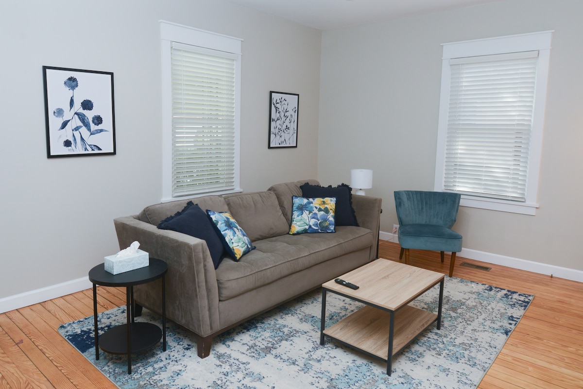 A cozy living room is showcased with a comfortable brown sofa adorned with decorative pillows. A coffee table sits in front, accompanied by a side table with a tissue box. Natural light streams through the white, partially open blinds, illuminating the soft area rug and wall art.