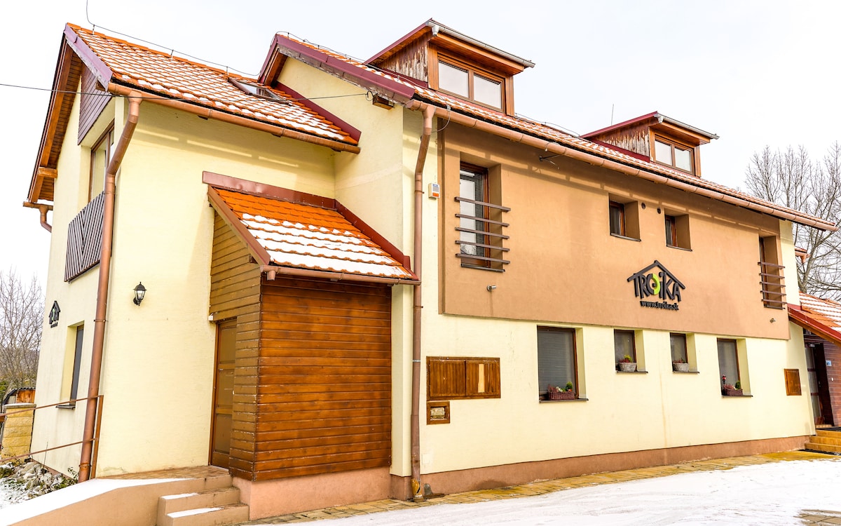 A three-story building is presented, featuring a combination of cream-colored and wooden facades. Windows are evenly spaced along the exterior, with some adorned with small flower pots. A sloped roof with red tiles provides a contrasting element against the winter setting.