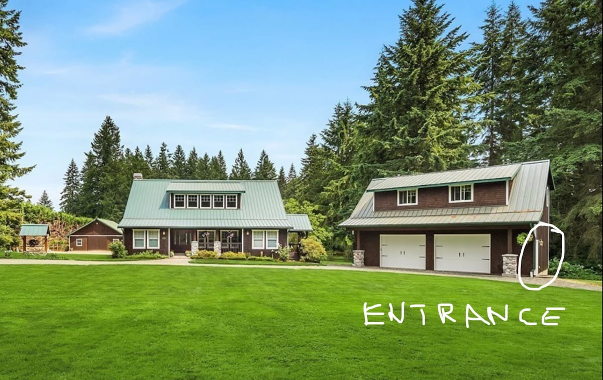 The exterior of the guesthouse is displayed, featuring a two-story building with a metal roof and large windows. A spacious lawn surrounds the property, providing an inviting entryway. A detached garage is present, complementing the rustic architectural style amidst a backdrop of lush trees.