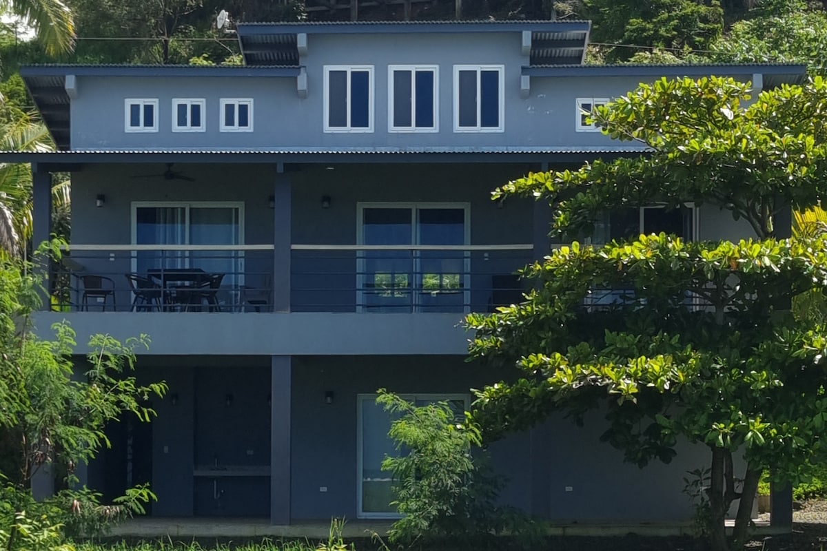 A modern two-story building is framed by lush greenery, featuring a large balcony with outdoor seating. The facade displays multiple windows, allowing natural light to enhance the interior space. The structure's neutral color complements the vibrant surrounding landscape.