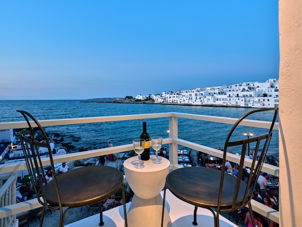 A balcony featuring two wrought-iron chairs and a small table holds a bottle of wine and two glasses. The view showcases the sea and whitewashed buildings along the shore, with the evening sky transitioning to dusk.